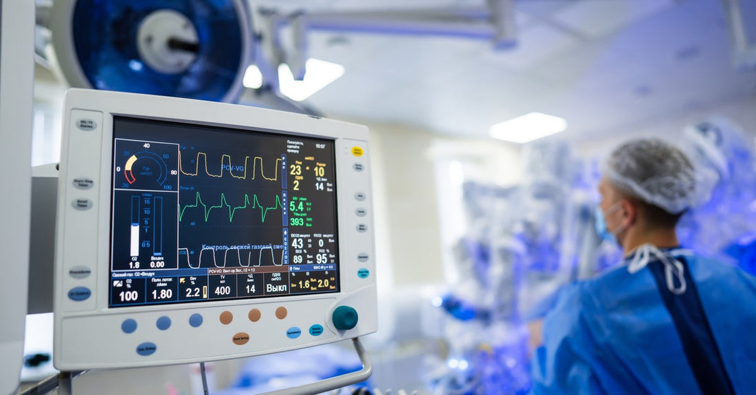 A masked person in surgical attire works beside a patient monitor showing vital signs and waveform readings.